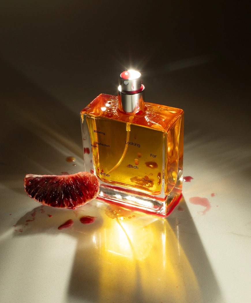 A 50ml bottle of Blinding Reflection from Tower Window by Looking for Each Other, glowing with amber botanical liquid beside a blood orange slice. A radiant image evoking the scent’s citrus-spice profile and plant-based purity—no synthetics, ever.
